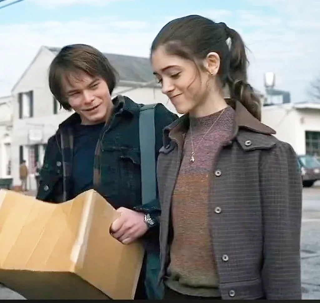 A young woman wears the Zing Apparel Stranger Things S01 Nancy Wheeler Purple Jacket while walking outdoors with a young man. Both are smiling, looking down at a large cardboard box, and chatting as they stroll along a street.